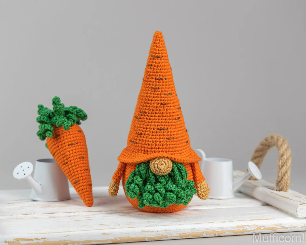 Crocheted carrot-shaped gnome decoration and a matching carrot topper, displayed on a white wooden surface with small white watering cans against a light gray background.