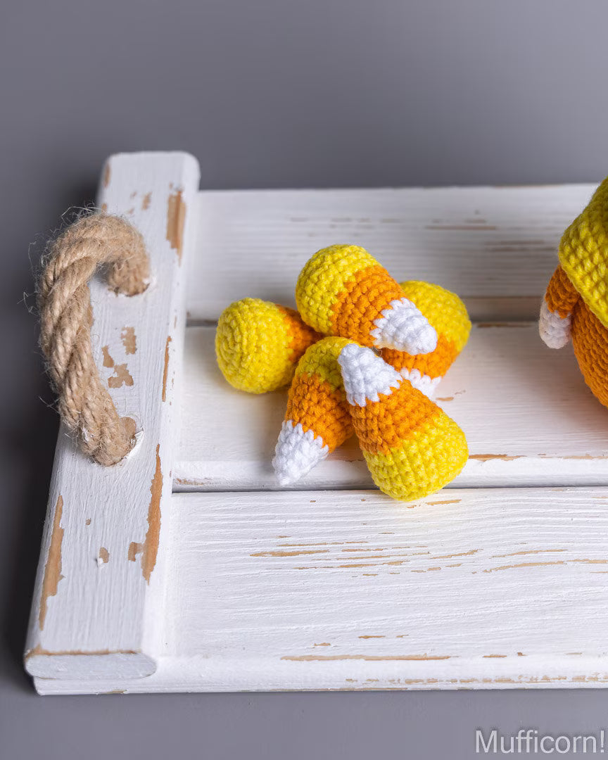 Crocheted candy corn toys arranged on a distressed white wooden tray with a rope handle, photographed against a smooth gray background. Part of another crocheted toy is visible on the right edge.