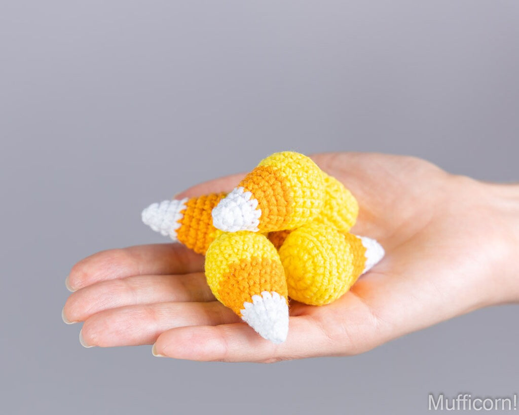 A hand holding four small crocheted candy corn toys in yellow, orange, and white against a plain gray background.