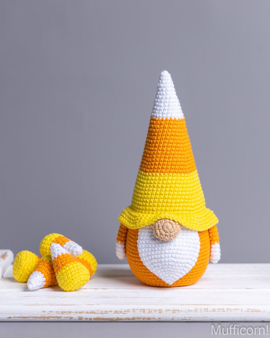 A crocheted candy corn gnome sits on a white wooden surface against a solid gray background, with several smaller loose candy corn pieces to its left.