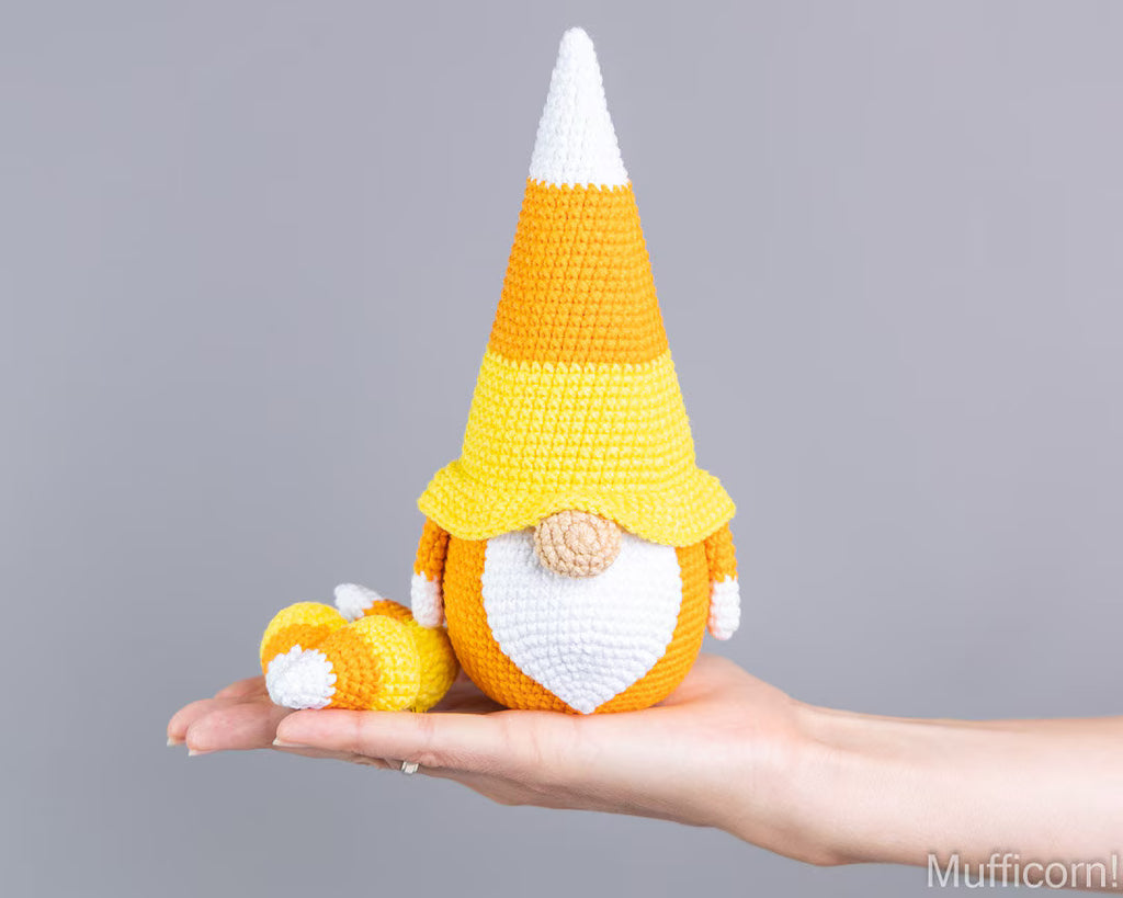 A crocheted candy corn gnome doll with a yellow, orange, and white striped hat and a white beard, being held in a person's hand against a plain gray background.