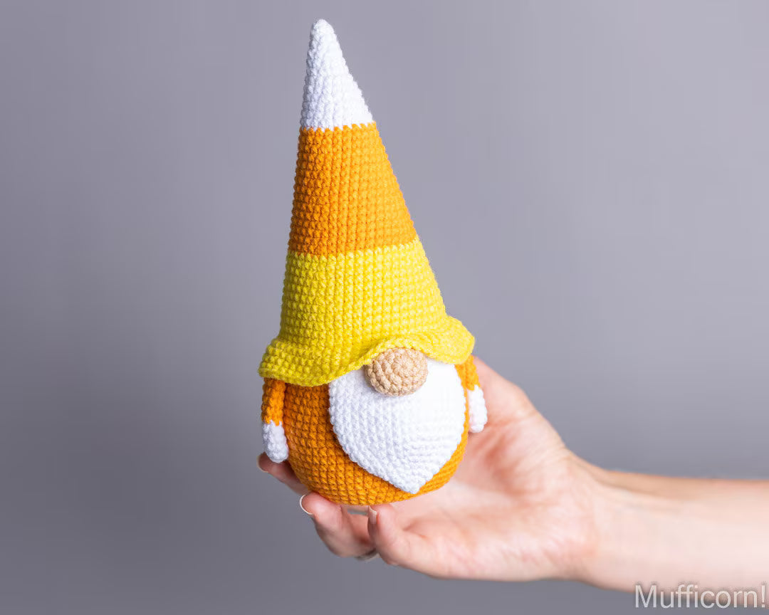 A crocheted candy corn gnome doll being held in a person's hand against a plain gray studio background.