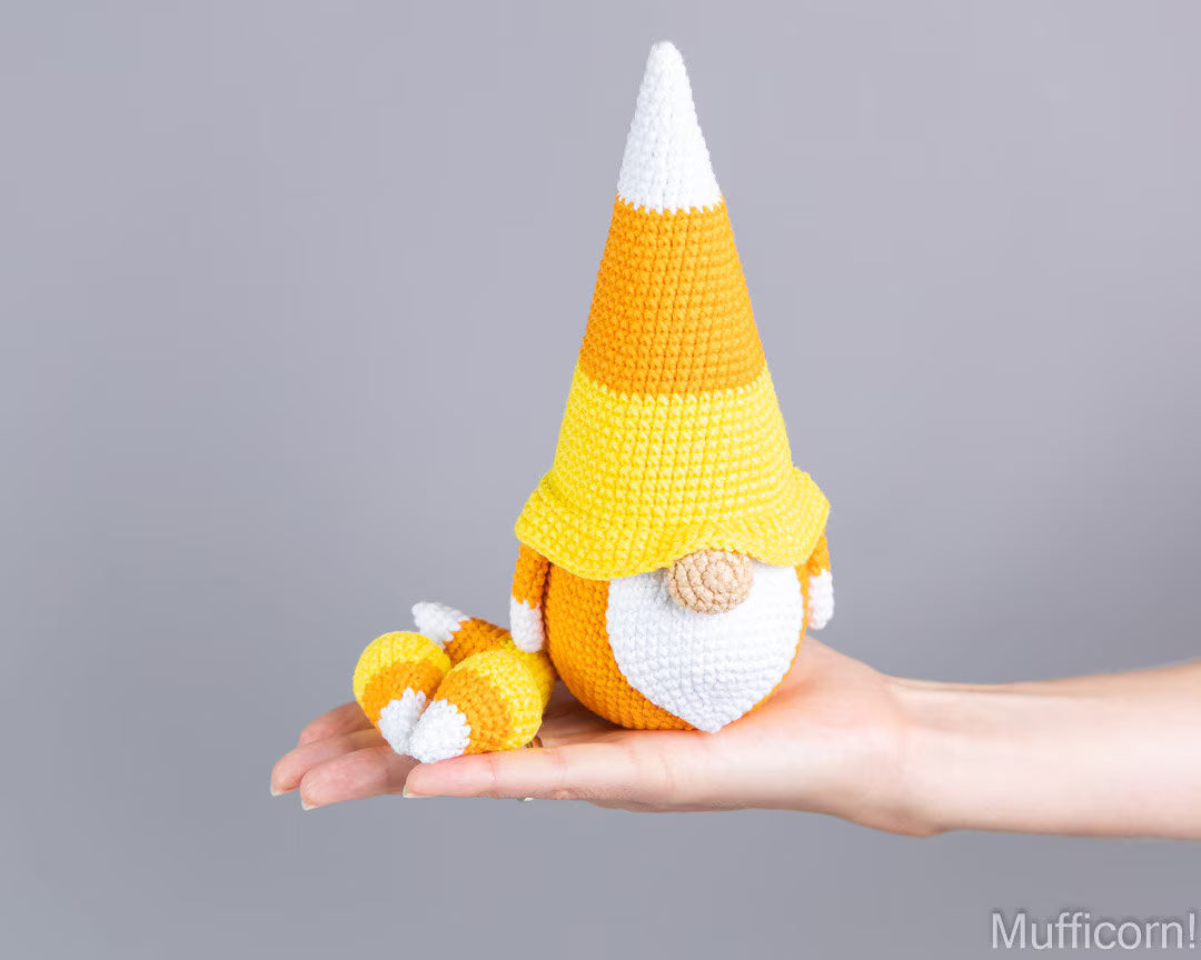 A crocheted candy corn gnome doll with a pointed yellow, orange, and white hat, white beard, and small matching candy corn accessory, displayed on an open hand against a plain gray background.