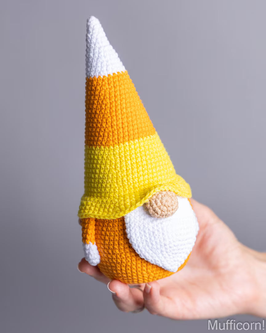 A crocheted candy corn gnome doll with a white beard and pointed hat, held in a person's hand against a plain gray background.