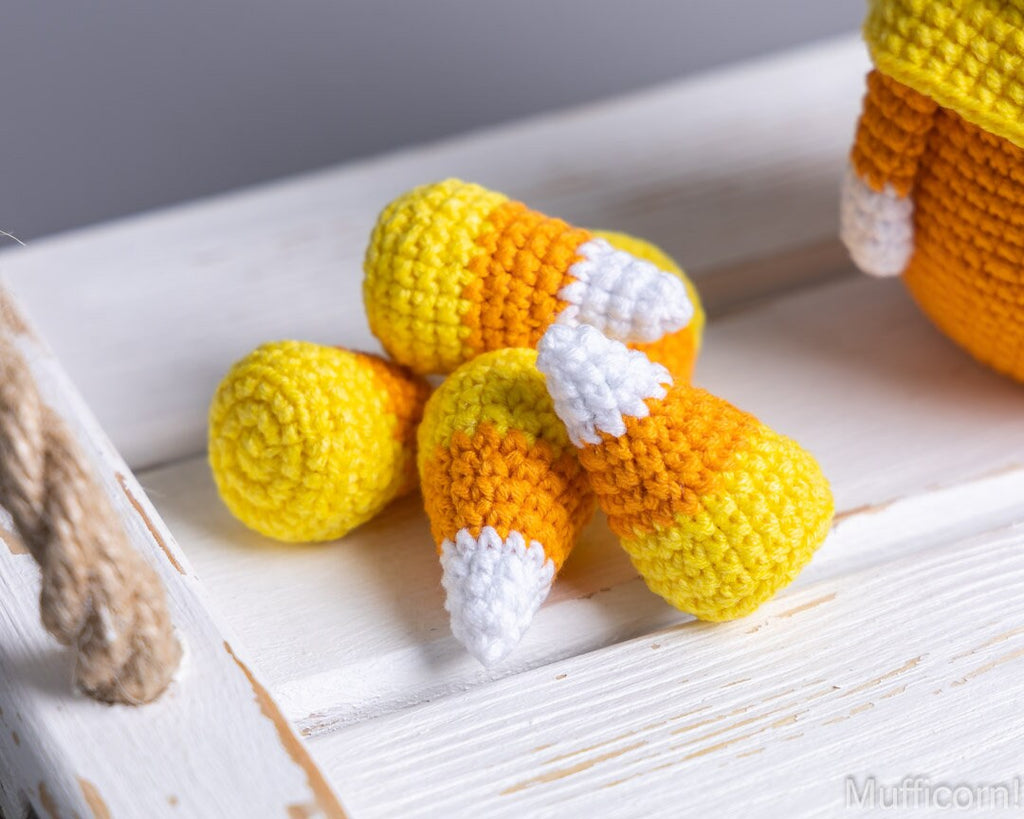 Three crocheted candy corn plush toys in yellow, orange, and white are arranged on a white wooden surface. A partial fourth candy corn is visible at the right edge, and a rope prop appears at the left edge.