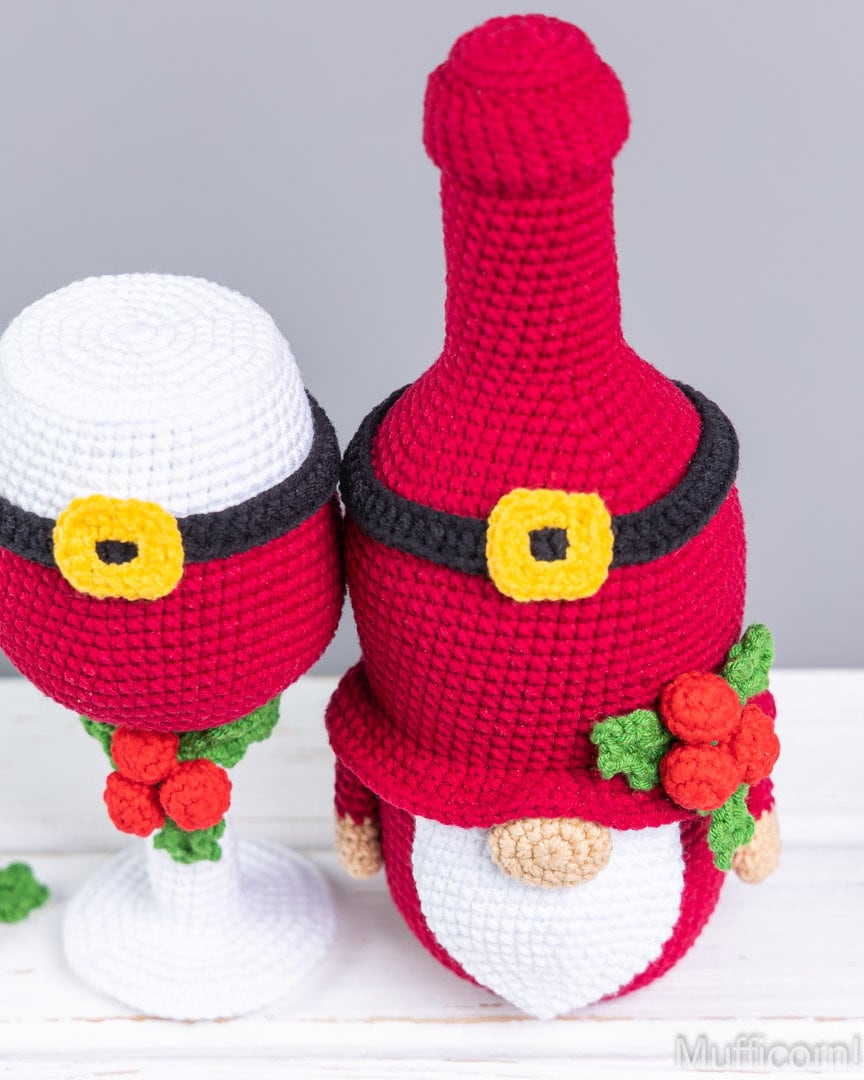 A crocheted Christmas-themed wine bottle cover and matching goblet, styled like Santa with a red body, black belt, yellow buckle, white beard, and red and green holly accents, set against a neutral gray background.