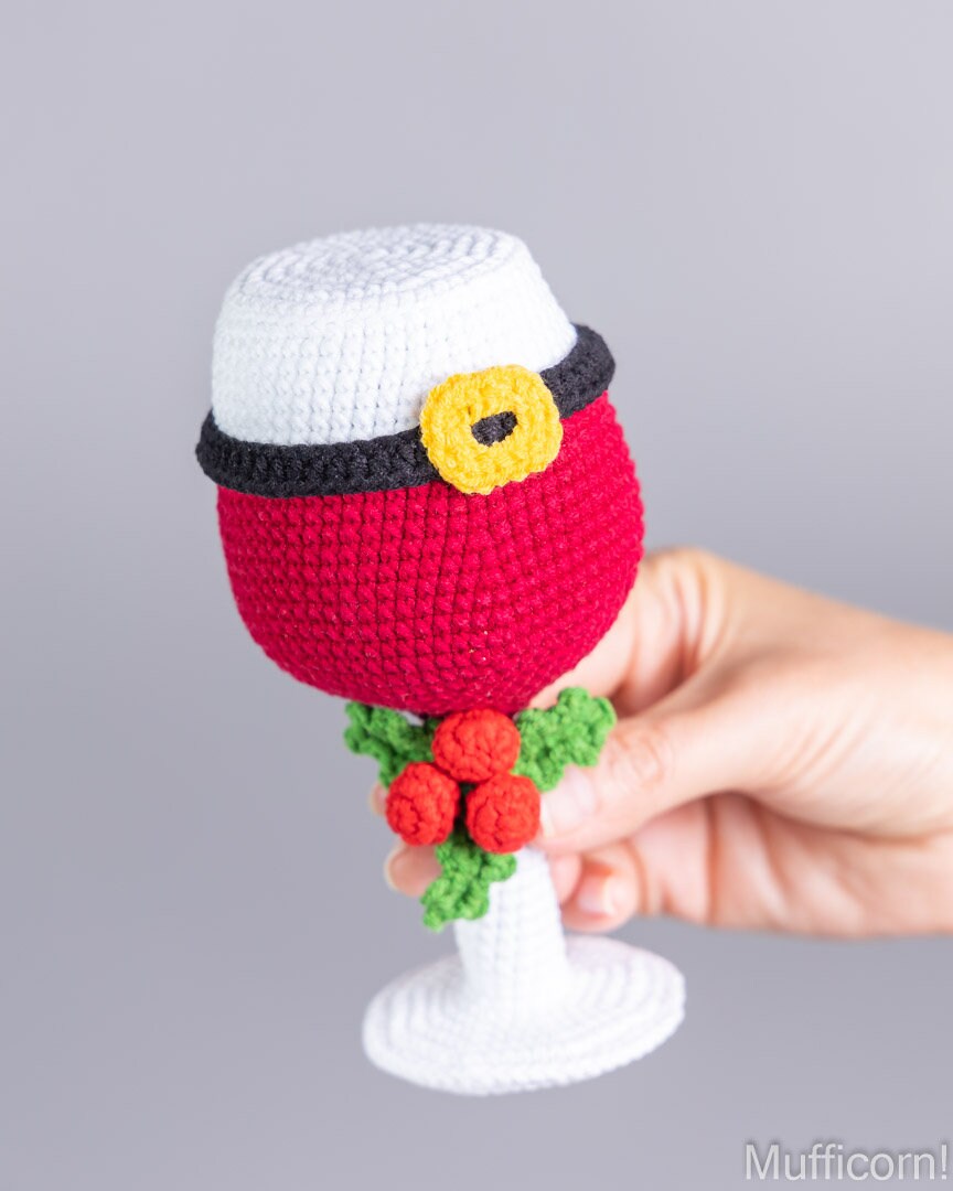 A crocheted Santa-themed wine glass held by a hand against a plain gray background. The crocheted design features a white Santa hat with a black band and yellow buckle, a red body, green leaves, and red berries on the stem.