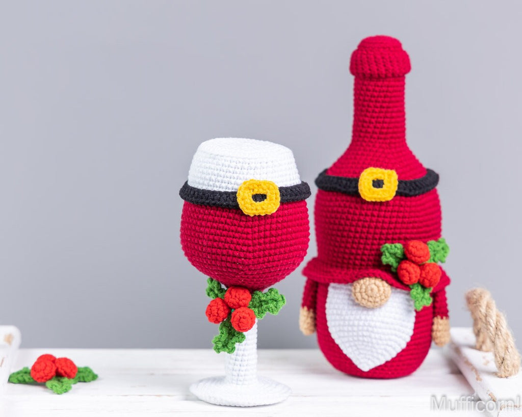 Two crocheted holiday-themed decorative items on a white surface against a light gray background: a red and white wine glass-shaped ornament with a black belt and yellow buckle, and a red and white gnome-shaped bottle cover with a white beard, red hat, and berry accents.