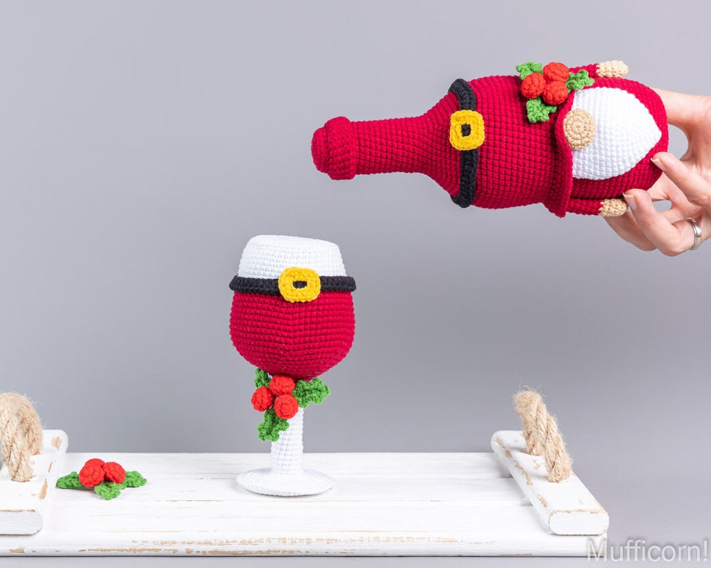 A crocheted Santa-themed wine bottle and matching wine glass are displayed on a white wooden surface against a light gray background. A human hand holds the crocheted Santa bottle in the upper right, while a completed matching set sits on the table.