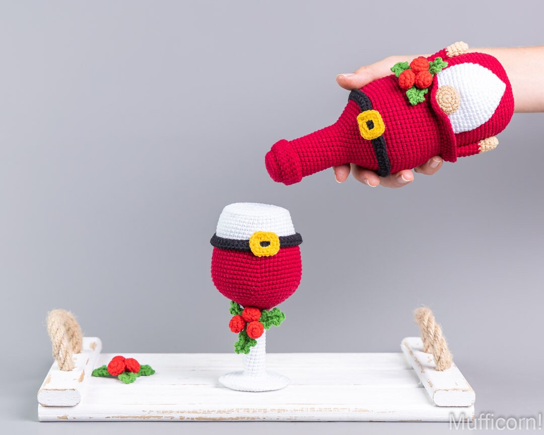 A crocheted Santa-themed wine glass cover and matching Santa glove are displayed on a white tray against a gray background. A hand holds the glove above the glass, which is decorated with red, white, black, yellow, and green details.