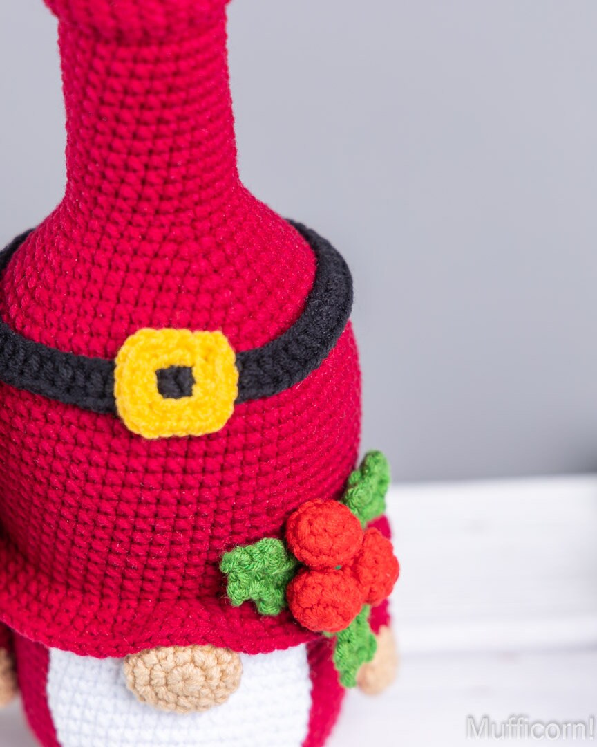 Close-up of a crocheted gnome figure in bright red, with a black belt, yellow buckle detail, white pants, and red-and-green flower accents. The background is a soft gray wall with a white surface below.