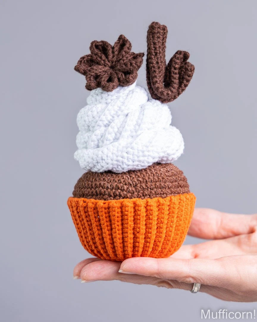 A crocheted cupcake toy held in a person's hand against a plain gray background. The cupcake has an orange base, a brown top, a white swirl of frosting, and brown decorative toppings.