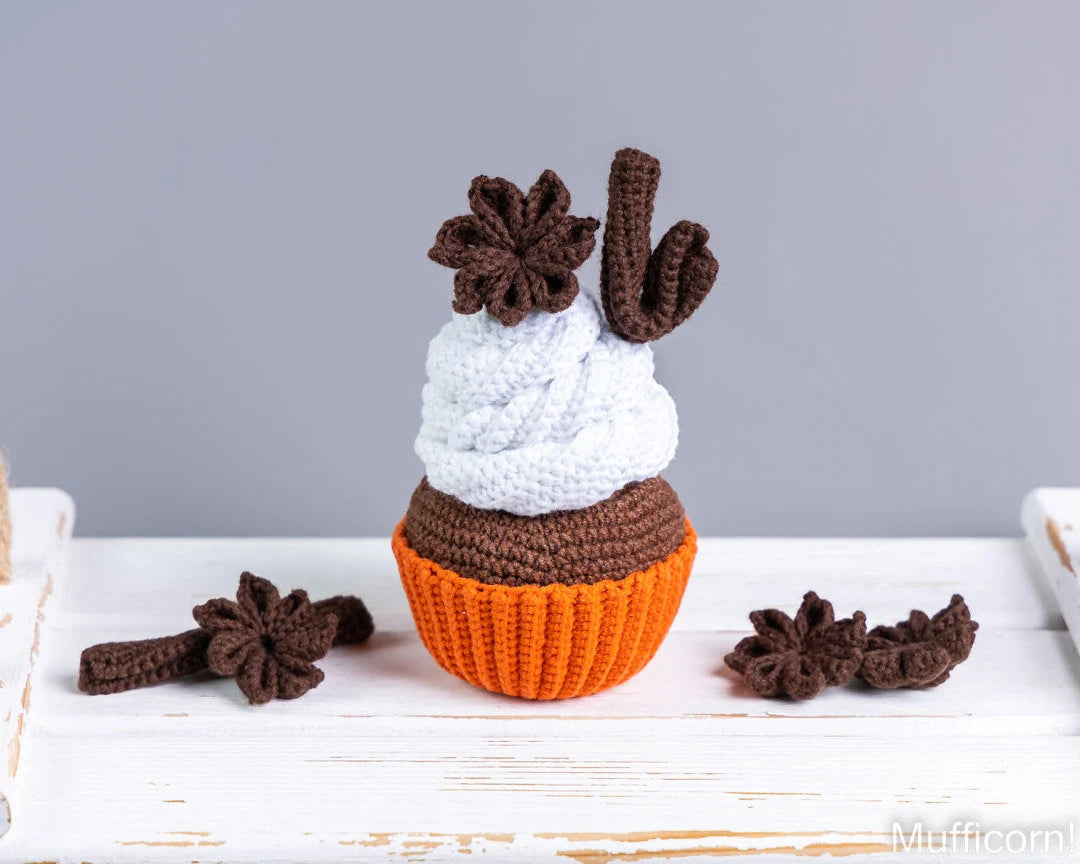 A crocheted cupcake decoration or doll with a white frosting top, brown cupcake body, orange wrapper, and brown star anise and cactus details, displayed on a white wooden surface against a gray background.