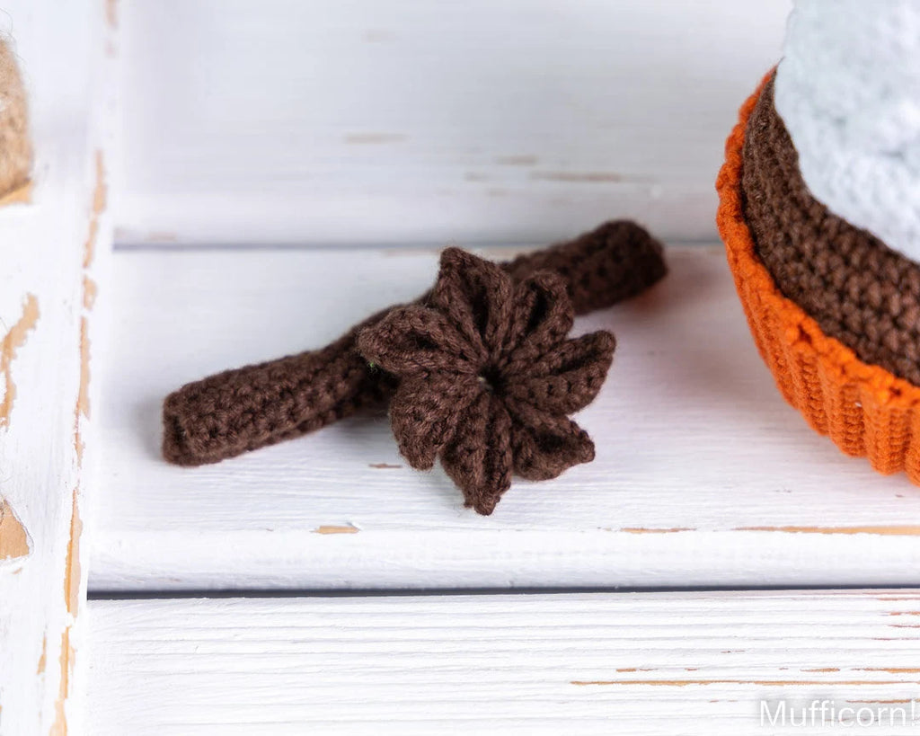 A dark brown crocheted headband with a matching flower accessory, displayed on a white wooden surface next to a partially visible orange and brown crocheted item.