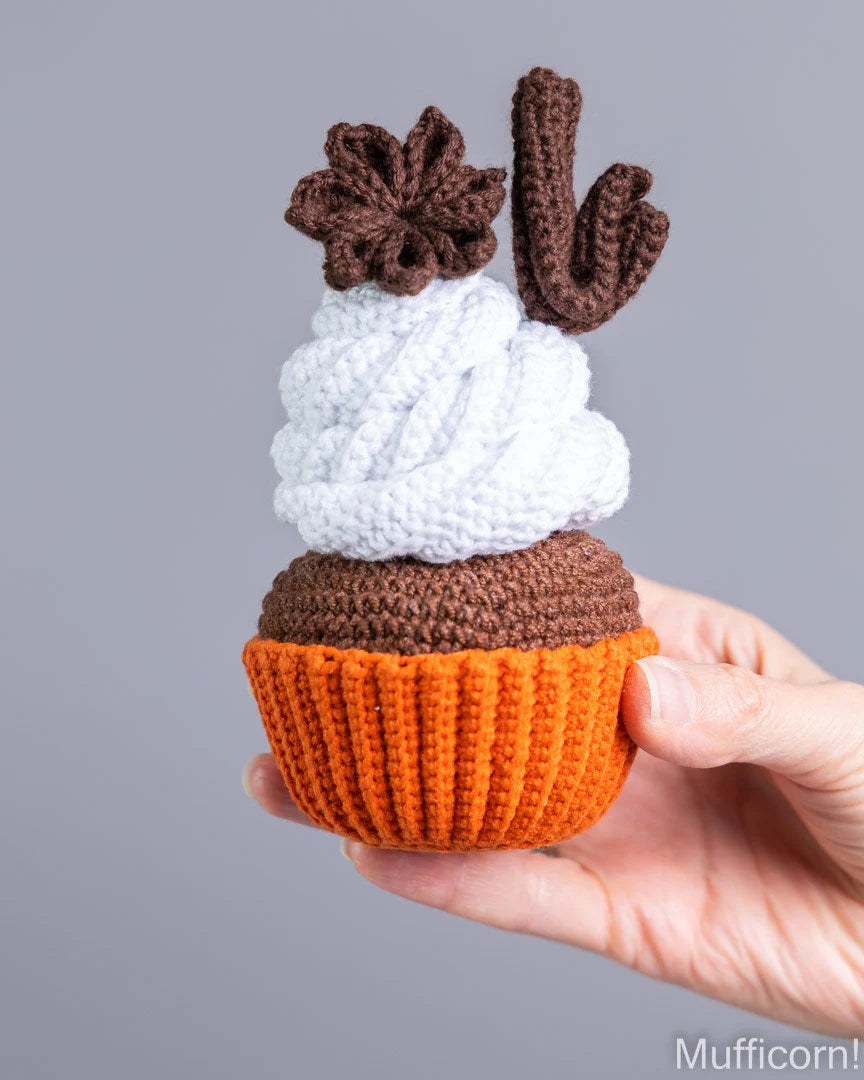 A hand holding a crocheted cupcake toy with an orange wrapper, brown frosting base, white frosting swirl, and brown decorative toppers against a plain gray background.