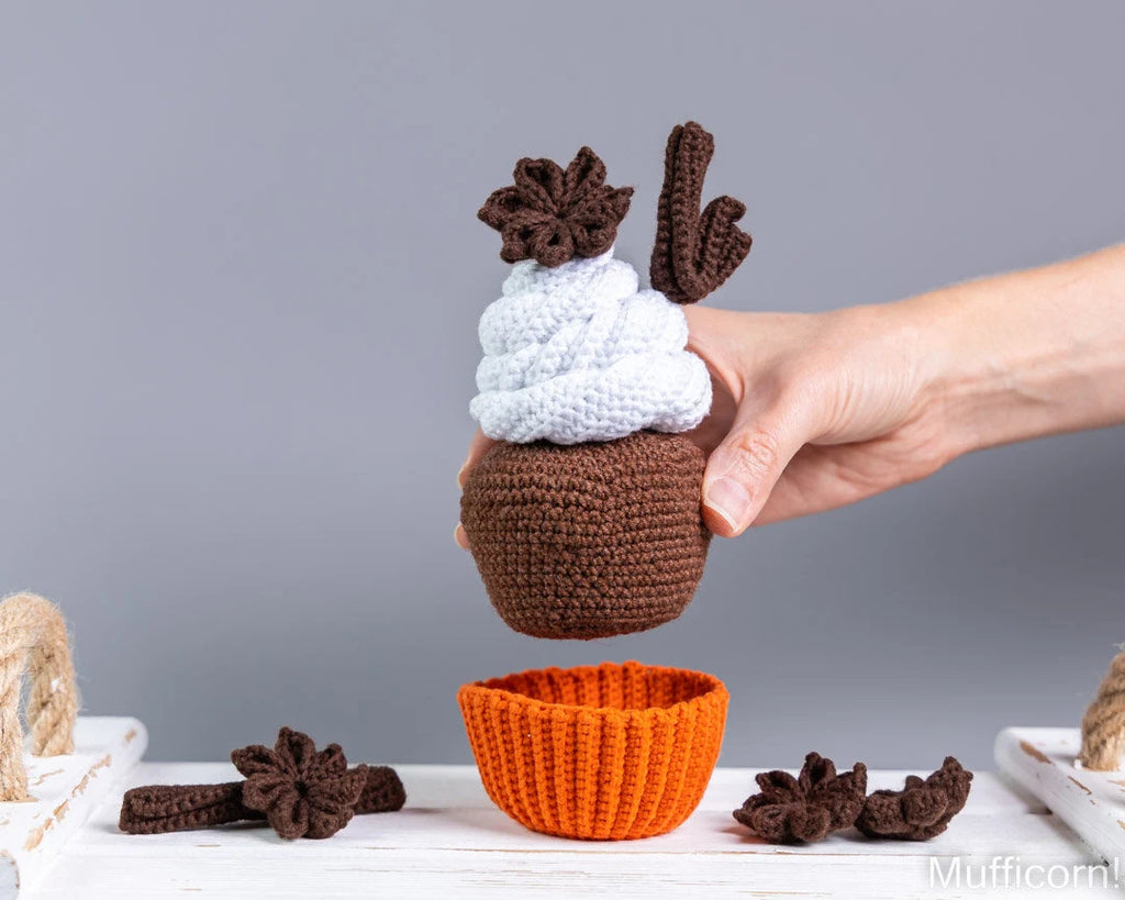A hand holds a crocheted acorn-shaped cup with a white frosting-like top and brown antlers, above an orange crocheted cup, on a white table with small brown decorative pieces against a gray background.