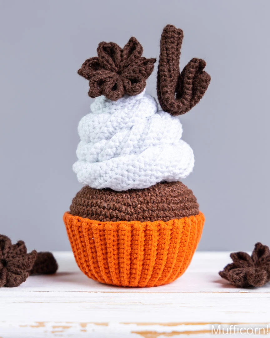 A crocheted cactus cupcake decoration with a white textured frosting top, brown cactus stem and flowers, and an orange cupcake liner base, displayed on a white rustic surface against a gray background.