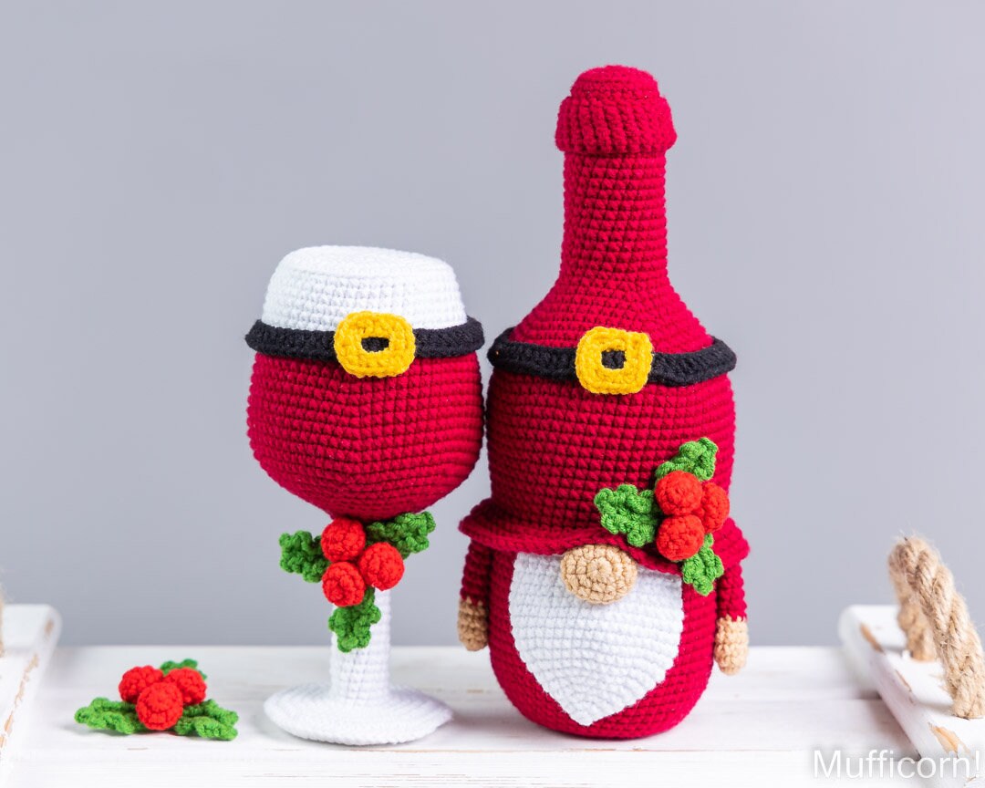 A crocheted Christmas gnome made from a red bottle and a white wine glass, both decorated with black belts, yellow buckles, white beards, red berries, and green holly leaves, displayed on a white surface against a gray background.