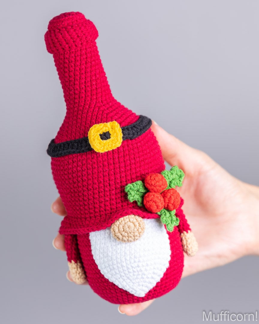 A crocheted Christmas gnome ornament shaped like a wine bottle, held in a hand against a plain gray background. The gnome has a red body, white beard, black belt with a yellow buckle, green leaves, and red berries.
