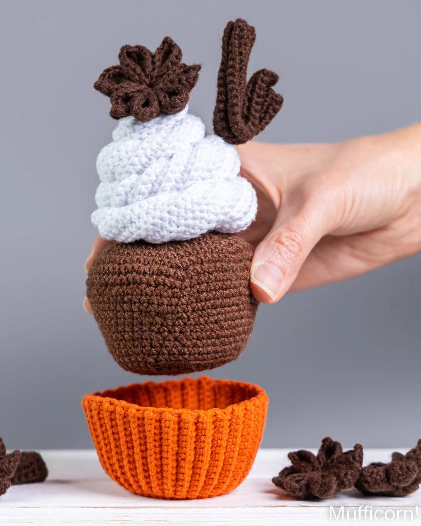 A crocheted cupcake toy being held by a person's hand, featuring a brown body, white frosting, and brown starburst decorations, with an orange cupcake liner below on a white surface against a gray background.