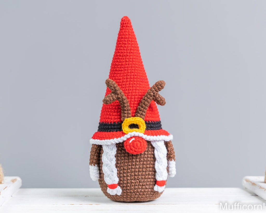 A crocheted Christmas gnome doll with a tall red hat, brown body, white braided scarf, and red nose, standing on a white surface against a light gray background.