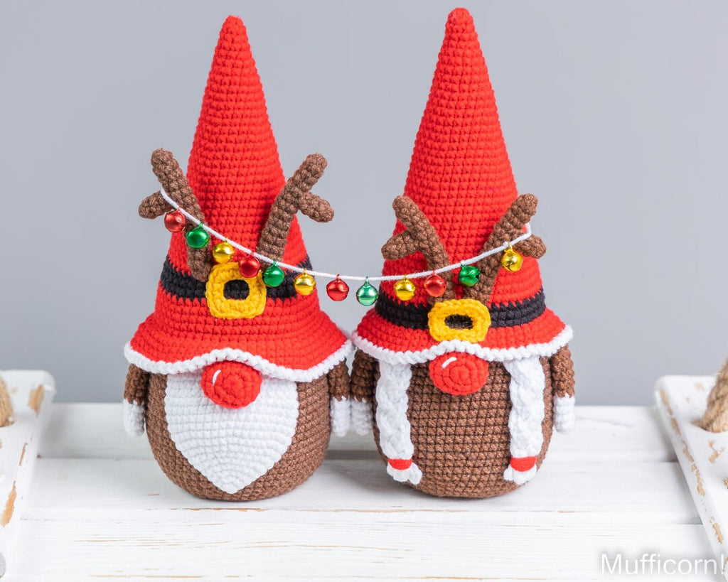 Two crocheted gnome holiday ornaments with red pointed hats, brown antlers, white beards, and colorful hanging ornaments, displayed on a white wooden surface against a light gray background.