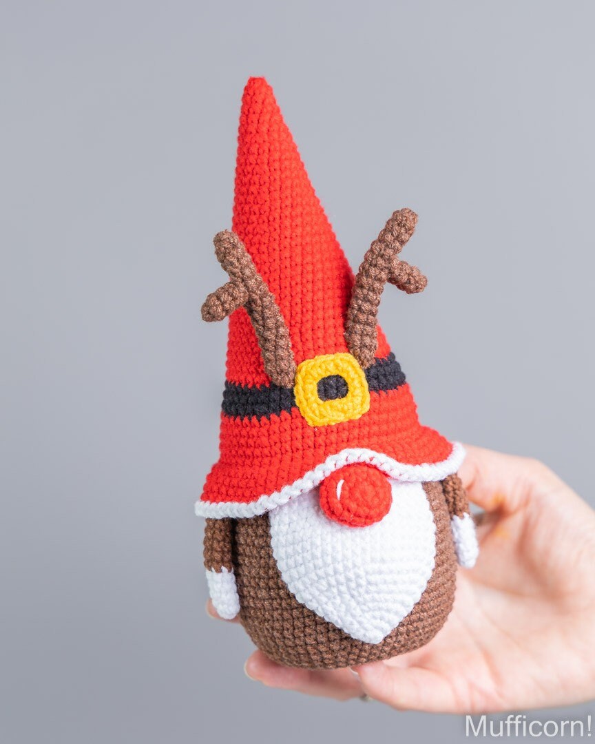 A crocheted Christmas gnome doll with a red pointed hat, brown antlers, white beard, and red nose, being held in a person's hand against a plain gray background.