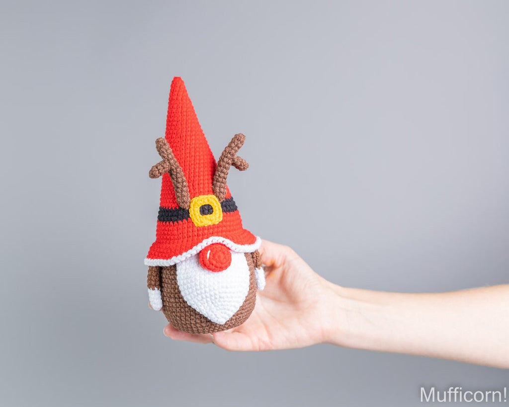 A hand holding a crocheted Christmas gnome doll with a red hat, brown antlers, white beard, and red nose against a plain gray background.