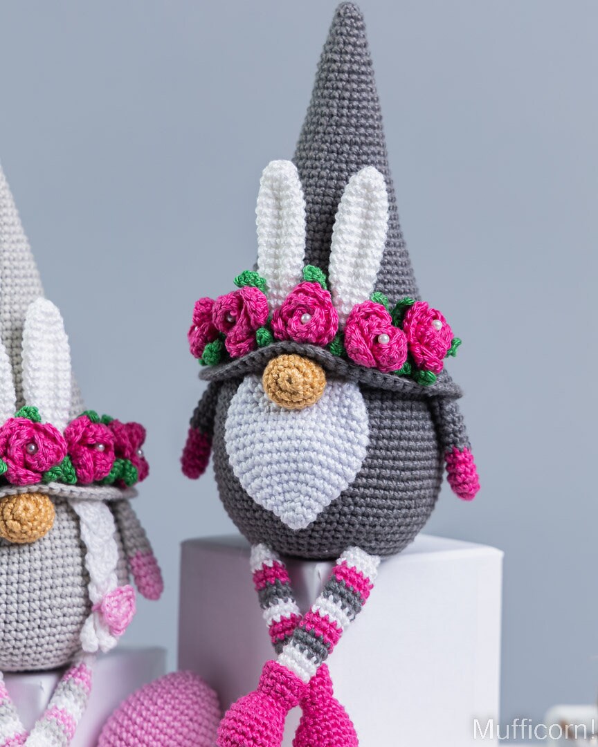 A crocheted bunny gnome doll with a gray pointed hat, white face and inner ears, pink flower crown, striped legs, and pink shoes, shown on a white pedestal against a light gray background.