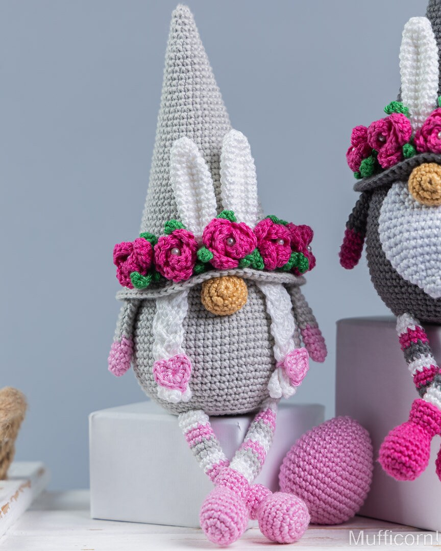 A crocheted bunny gnomelike doll with a tall gray hat, white ears, a pink flower crown, and pink-and-white striped legs, posed on a light gray surface with a matching pink egg nearby. A second similar doll is partially visible on the right.