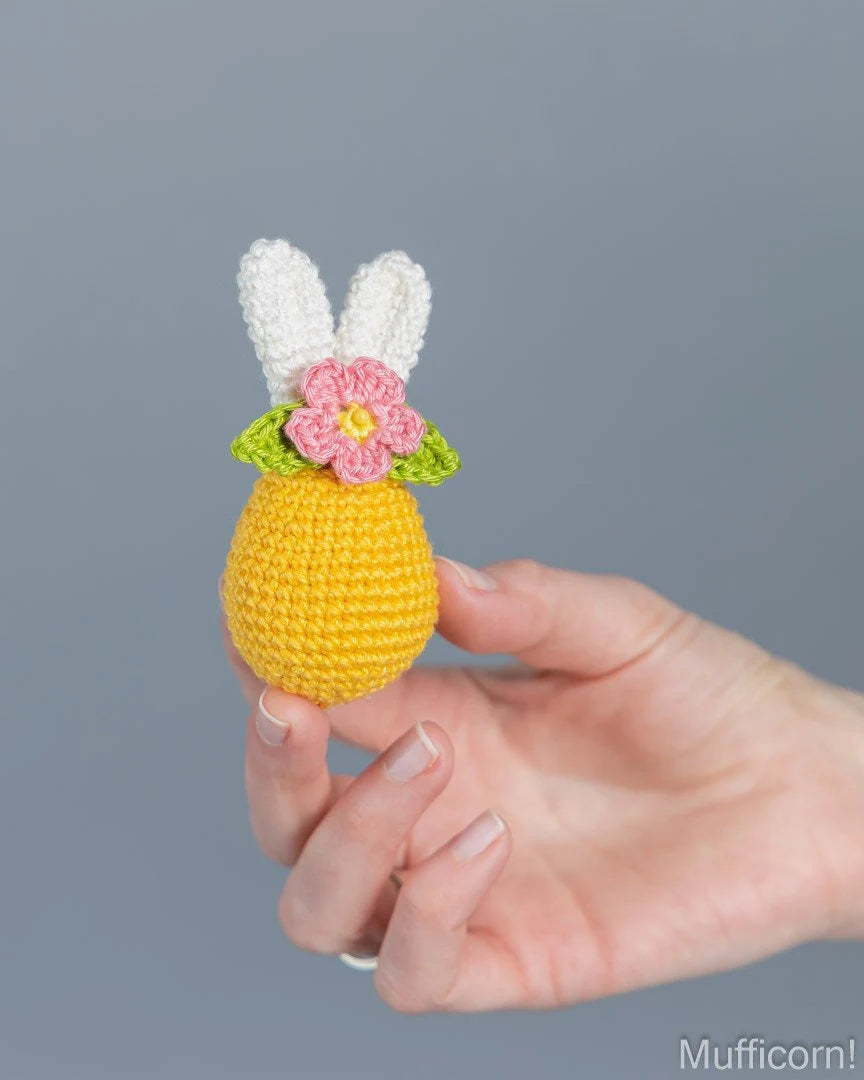 A hand holding a small crocheted bunny-shaped pineapple in yellow, with white bunny ears, green leaves, and a pink flower on top, against a plain gray background.