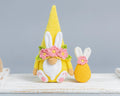 A crocheted Easter gnome doll in bright yellow with white bunny ears, a white beard, and pink flowers, shown next to a smaller matching crocheted egg on a white wooden surface against a soft gray background.