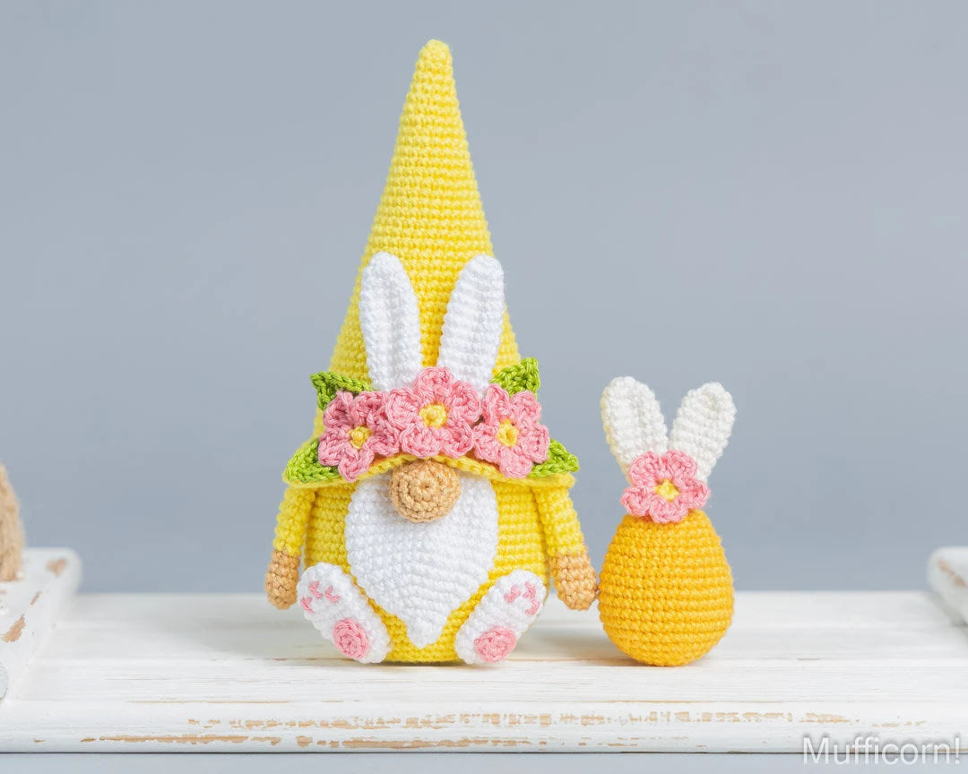 A crocheted Easter gnome doll in bright yellow with white bunny ears, a white beard, and pink flowers, shown next to a smaller matching crocheted egg on a white wooden surface against a soft gray background.