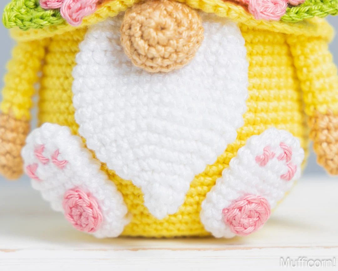 Close-up of a crocheted amigurumi-style character or plush toy, showing a bright yellow body, large white belly patch, pink paw pads, tan ears/feet, and floral trim at the top.