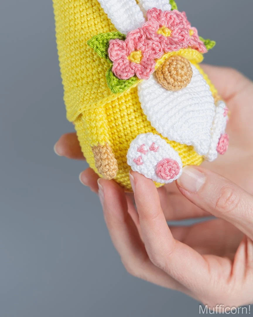 A close-up of a hand holding a crocheted bunny-shaped item, likely a pouch or small accessory, in bright yellow with white, pink, green, and tan details. The background is a clean, neutral gray.