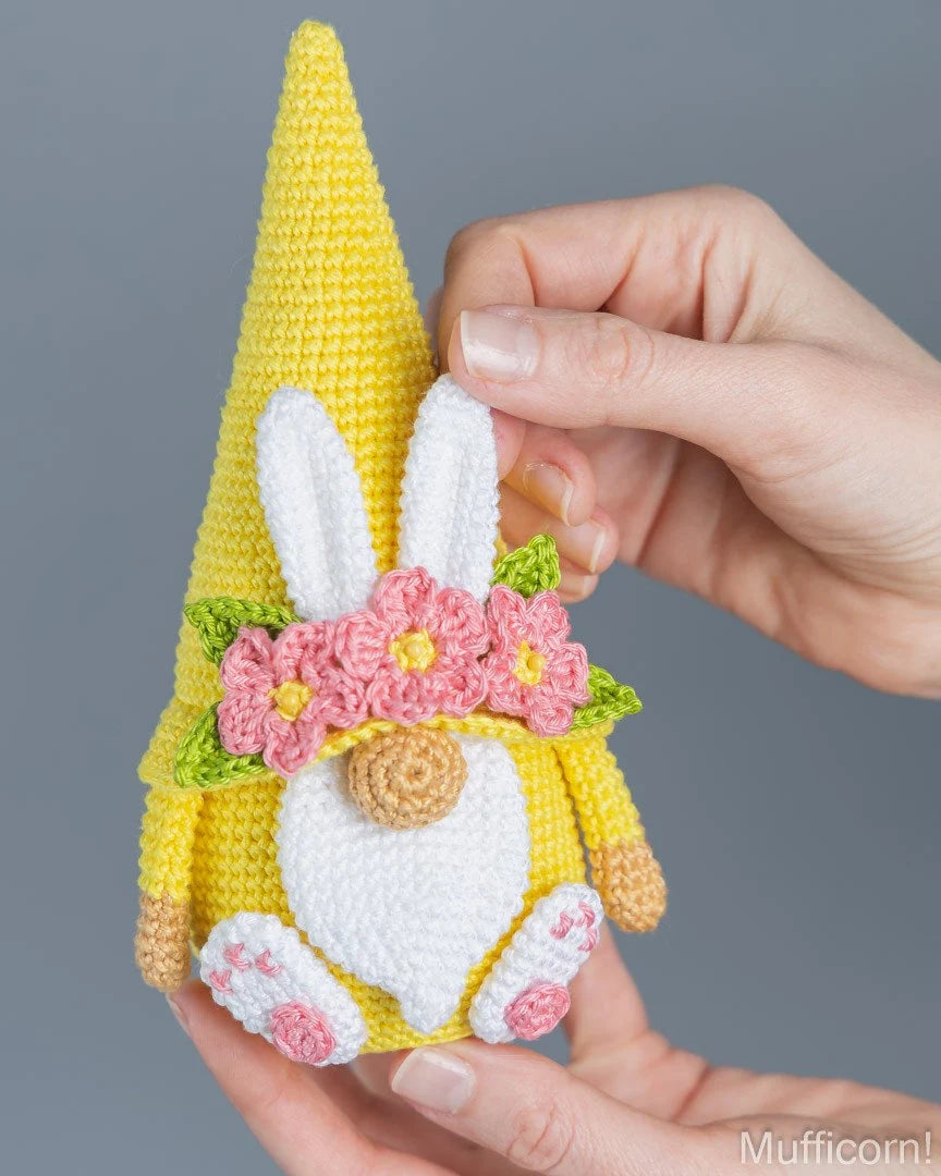 A crocheted Easter gnome doll with a yellow hat, white bunny ears, pink flowers, and a white beard, held by two hands against a solid gray background.