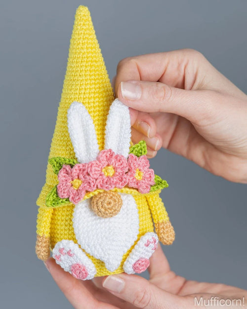 A crocheted Easter gnome doll with a yellow hat, white bunny ears, pink flowers, and a white beard, held by two hands against a solid gray background.