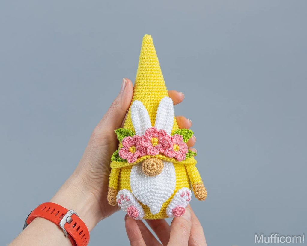 A crocheted Easter gnome doll with a yellow hat and body, white beard and bunny ears, pink flowers, and green leaves, held in two hands against a solid gray background.