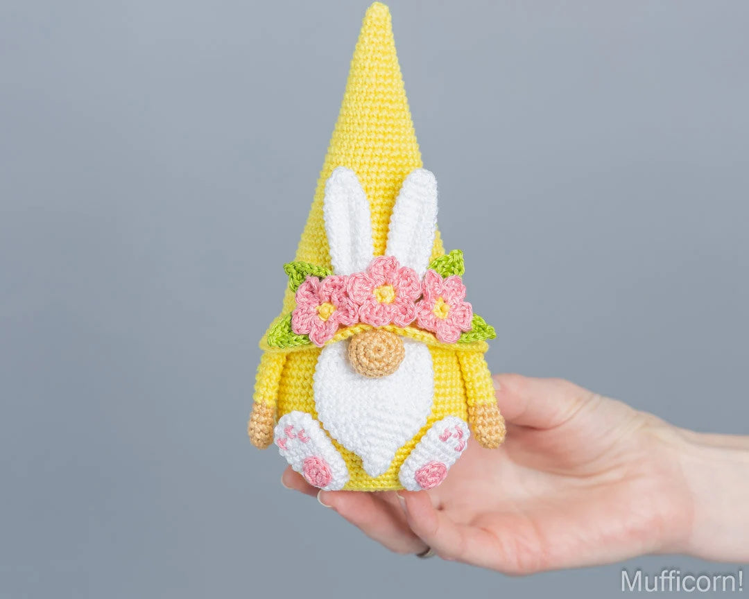 A crocheted bunny gnome with a yellow pointed hat, white body, pink flowers, and green leaves, held in a person's hand against a plain gray background.