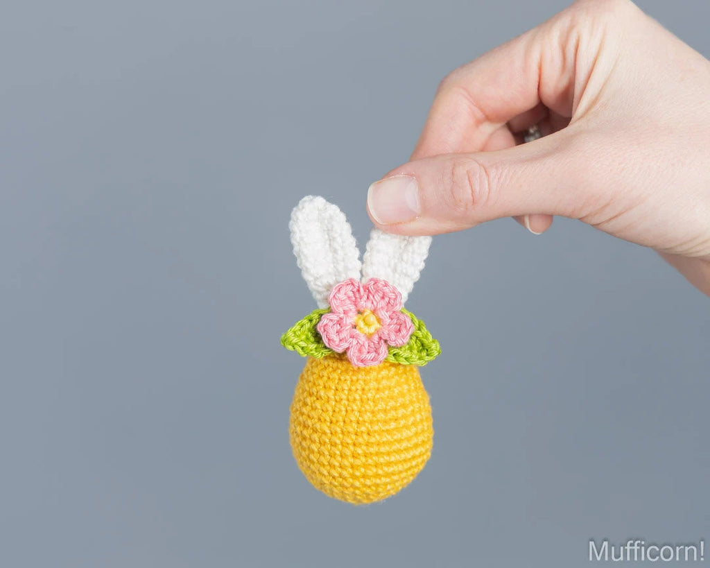 A hand holds a small crocheted bunny egg ornament against a solid gray background. The ornament features a yellow body, white bunny ears, green leaves, and a pink flower with a yellow center.