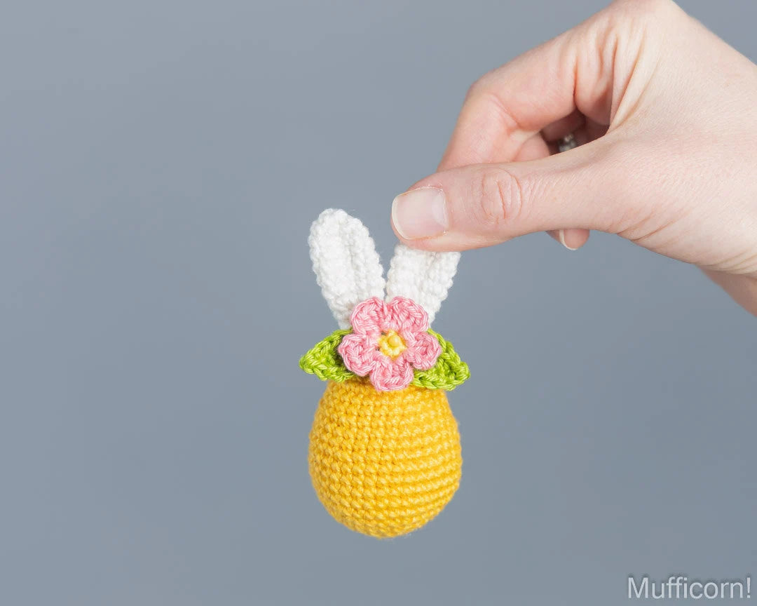 A hand holds a small crocheted bunny egg ornament against a solid gray background. The ornament features a yellow body, white bunny ears, green leaves, and a pink flower with a yellow center.