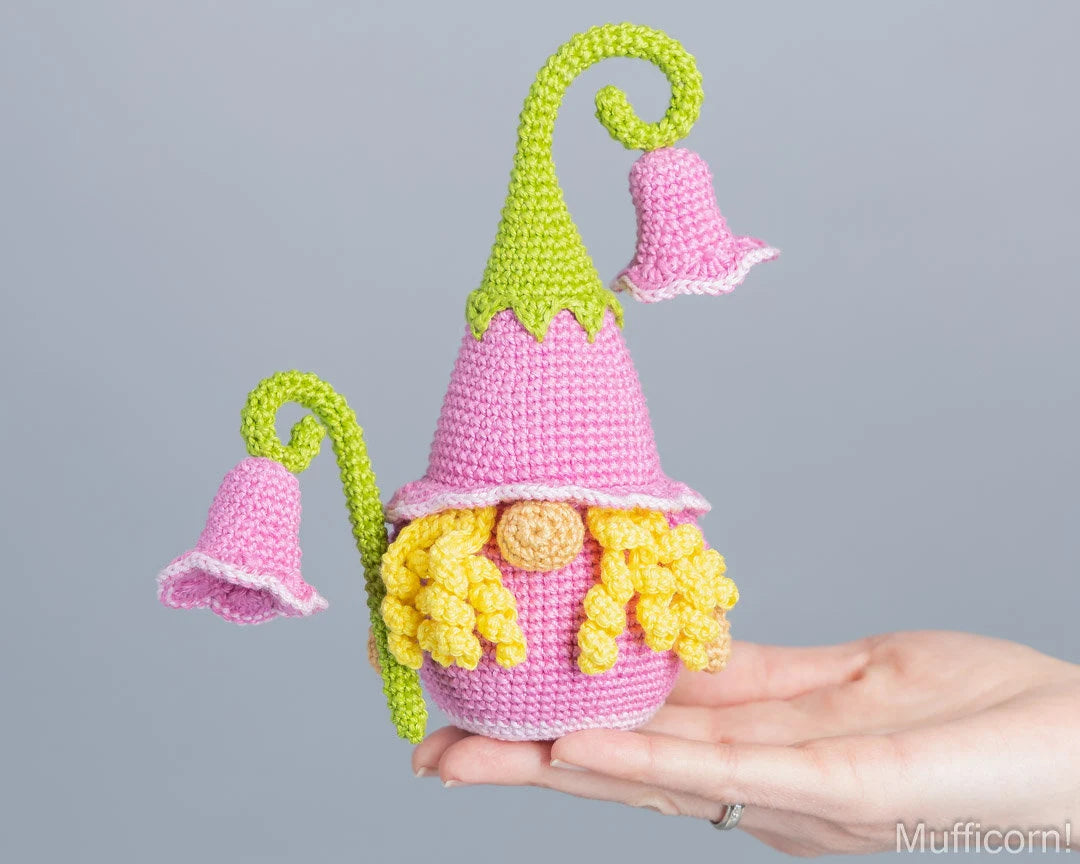 A crocheted gnome doll with campanula and a pink hat held in a person's hand against a plain gray background.