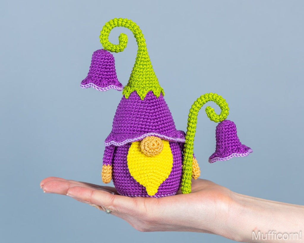 A crocheted gnome figurine with a purple hat and bell, yellow face and chest, and green curled stems, displayed on a person's open hand against a plain light blue-gray background.