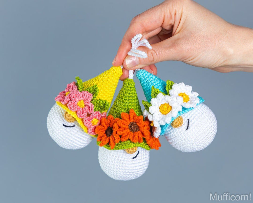 A hand holds three small crocheted gnome figurines wearing colorful party hats with flowers, against a plain gray background. The watermark 'Mufficorn' is visible in the bottom right corner.