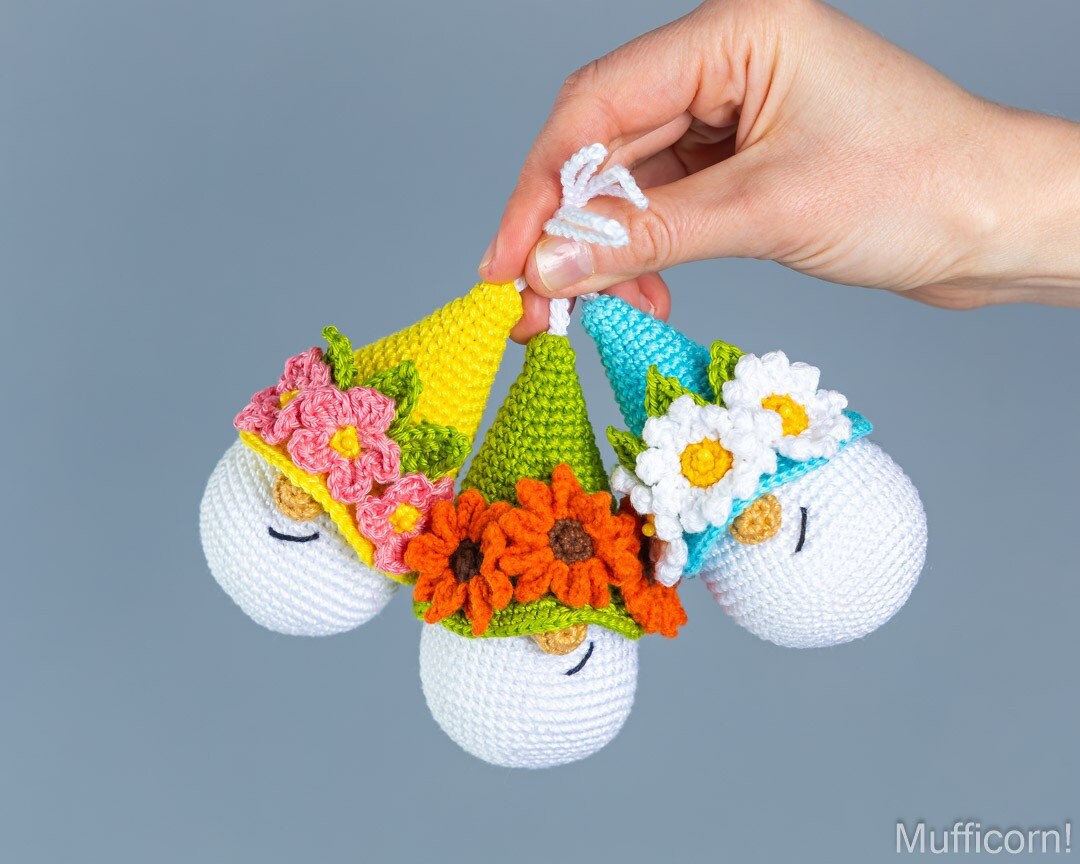 A hand holds three small crocheted gnome figurines wearing colorful party hats with flowers, against a plain gray background. The watermark 'Mufficorn' is visible in the bottom right corner.