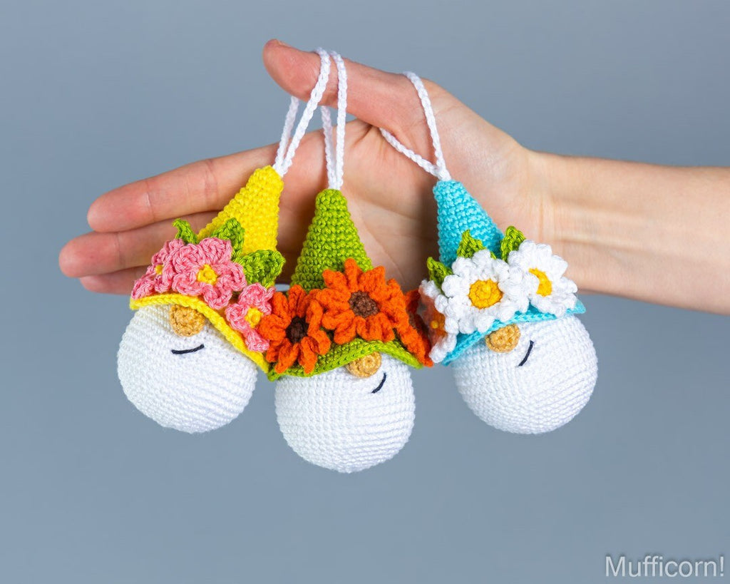 Three crocheted hanging ornaments shaped like sleeping white eggs with colorful pointed hats and flower crowns, held in a person's hand against a plain gray background.