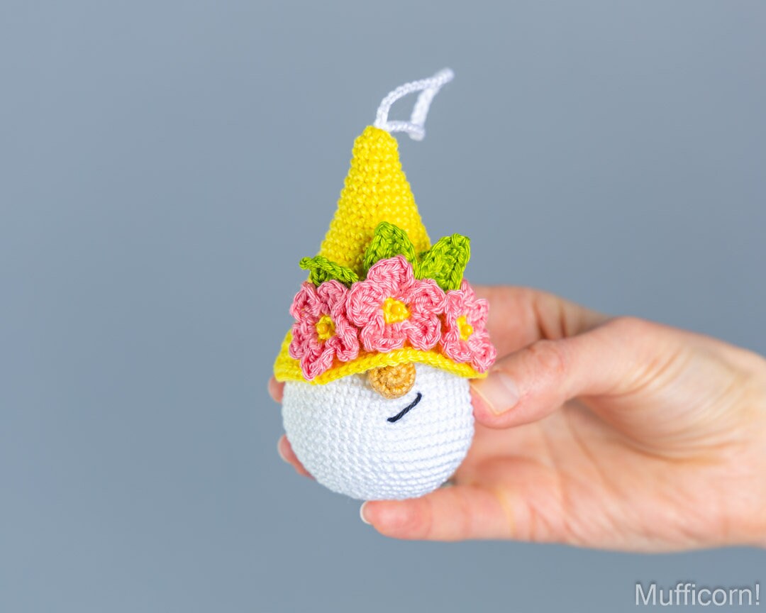 A hand holding a crocheted gnome ornament wearing a yellow hat with pink flowers and green leaves, against a plain gray background.