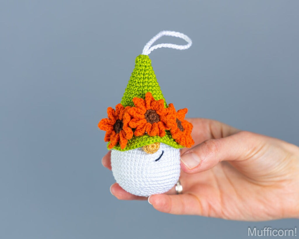 A hand holds a small crocheted gnome ornament against a solid gray background. The gnome has a white body, a green pointed hat, and orange flowers on its head.