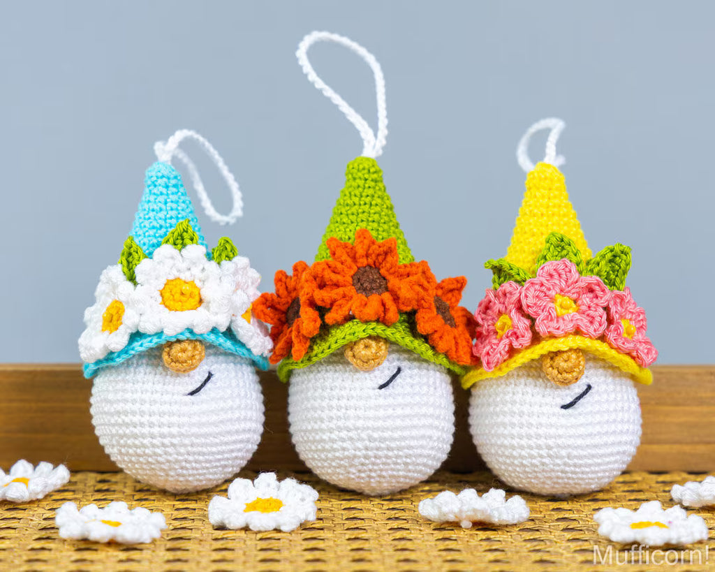 Three crocheted gnome ornaments with colorful pointed hats and flower crowns, displayed on a woven mat against a gray background.