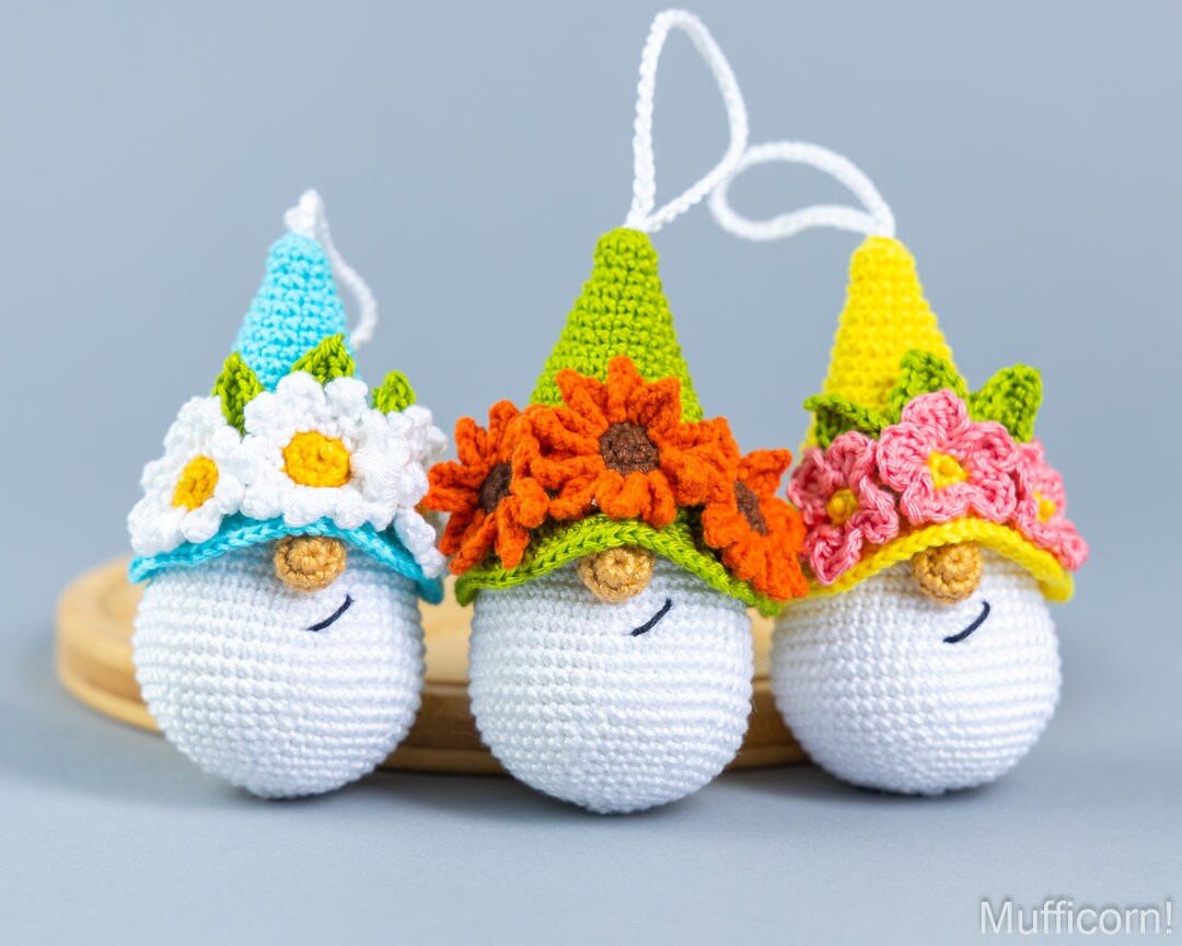 Three crocheted gnome ornaments wearing colorful flower hats, arranged on a small wooden tray against a light gray background. The ornaments have white bodies, embroidered black eyes and smiles, and are topped with pointed hats in blue, green, and yellow decorated with flowers.